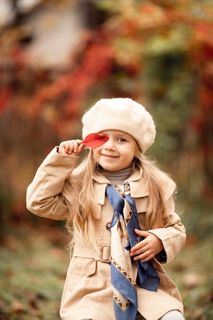 Pretty funny little blonde girl in a beret and coat holds a red leaf of a tree cheerful emotions The child plays in the fall in nature