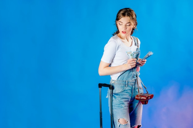 Красивая девушка лет 20 в джинсовом комбинезоне с испуганным выражением на лице позирует на синем фоне с чемоданом и деньгами в руках