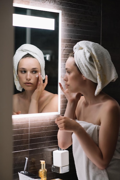 Foto bella ragazza che applica crema sul viso mentre si guarda il riflesso speculare in un bagno accogliente