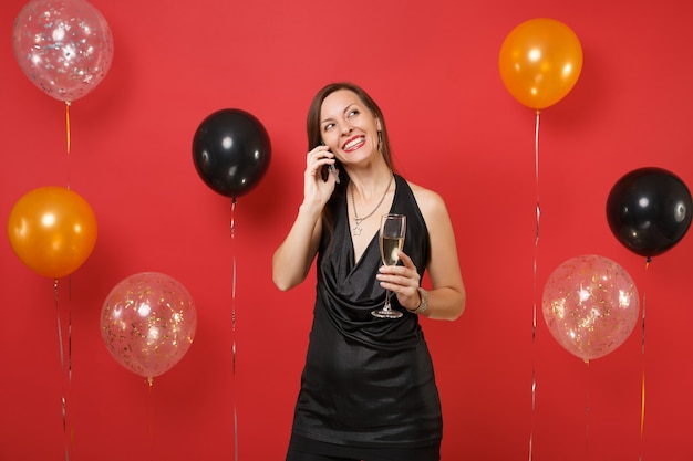 Pretty girl in black dress holding glass of champagne talk on mobile phone conducting pleasant conversation on bright red background air balloons. Happy New Year birthday mockup holiday party concept.