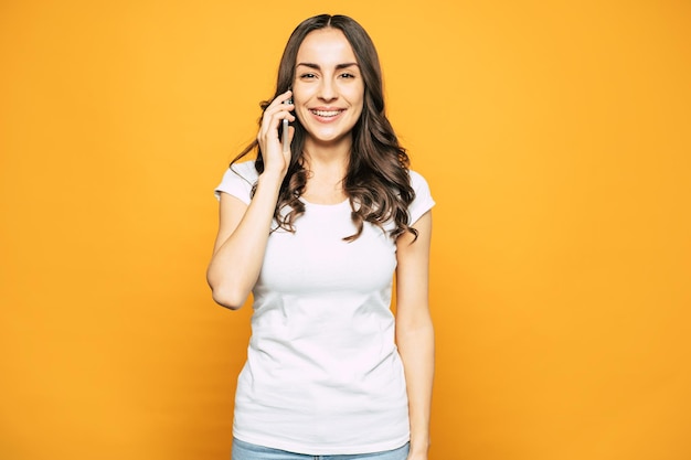 Pretty girl in casual outfit of T-shirt and denim jeans is talking to her boyfriend by mobile phone next to spice orange wall