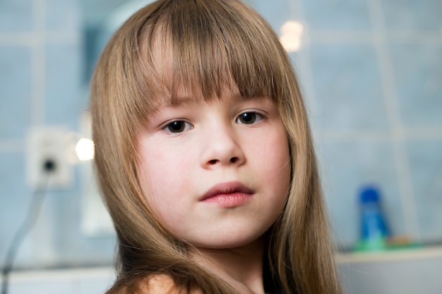 Pretty girl face portrait, child with beautiful eyes and long wet fair hair on blurred background of bathroom.