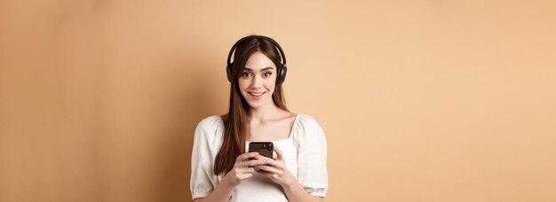 音楽を聴くカメラで笑顔と携帯電話アプリ ベージュ backgro を使用してヘッドフォンでかわいい女の子