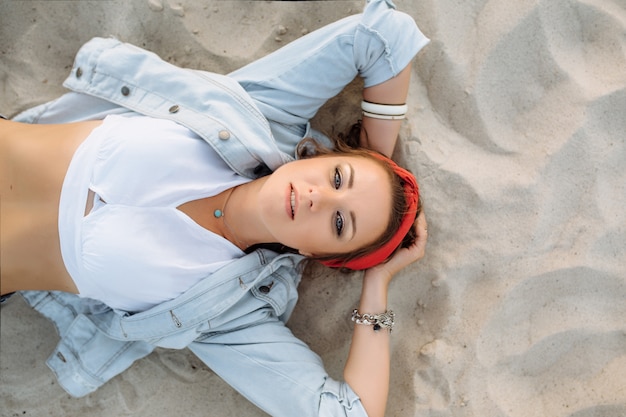 A pretty girl in jeans, short shorts, a red hoop and a white bra is sitting on the sand