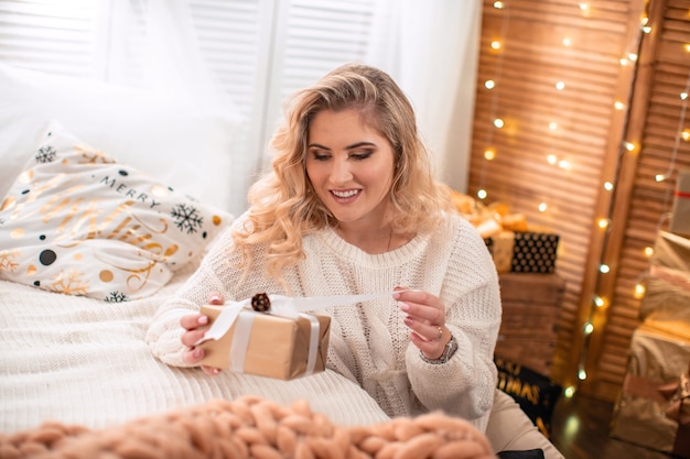 A pretty girl in a light evening makeup, sitting on the floor by the bed, smiling opens a box with a gift. Christmas gift, happy moment, women's happiness