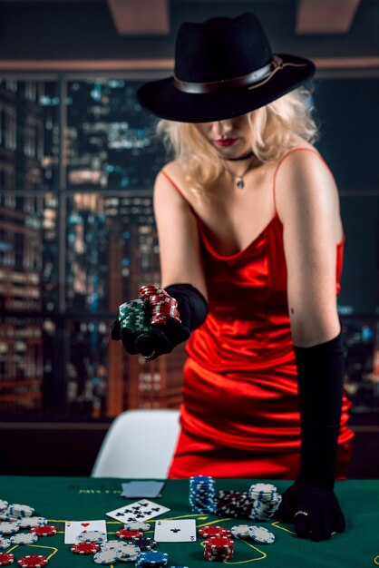 Pretty girl in red shiny dress and hat playing poker at casino