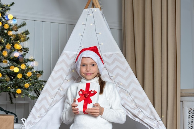 A pretty girl in a Santa hat holds her gifts for Christmas New Year lying in a children's room decorated with garlands