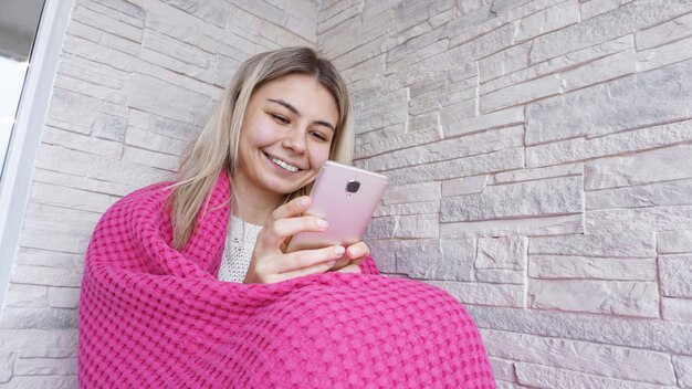 Красивая девушка сидит на подоконнике со смартфоном в руках. У нее длинные светлые волосы, она улыбается и смотрит в свой телефон.