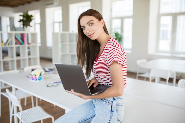 Bella ragazza in maglietta a righe appoggiata sulla scrivania che guarda sognante nella fotocamera mentre tiene il laptop in mano che lavora in un ufficio moderno