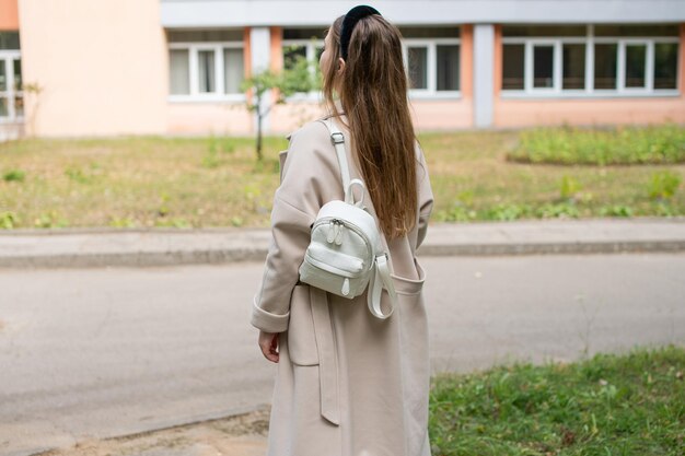 Симпатичная студентка в пальто с чашкой кофе гуляет возле учебного заведения