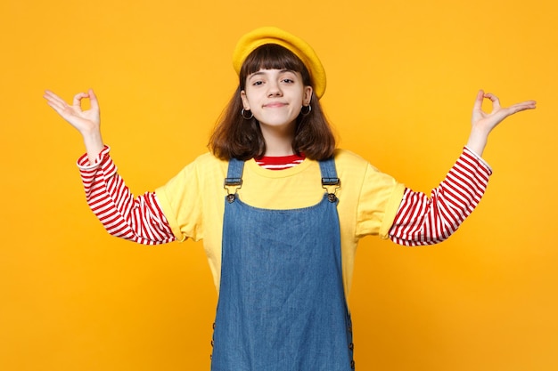 Pretty girl teenager in french beret, denim sundress hold hands in yoga gesture, relaxing meditating isolated on yellow wall background. People sincere emotions, lifestyle concept. Mock up copy space.