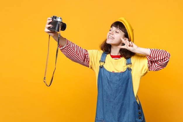 Pretty girl teenager in french beret doing selfie shot on retro vintage photo camera, showing victory sign isolated on yellow background. People sincere emotions lifestyle concept. Mock up copy space.