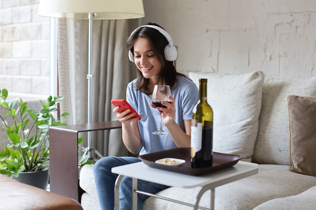 Pretty girl using her smartphone on couch at home in the living room. Listening music, drinking red wine, relaxation after a hard week at work.