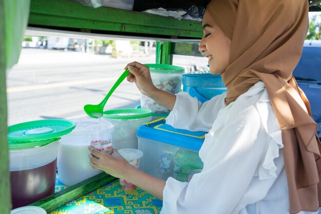 A pretty girl in a veil sells es campur using a scoop to get coconut milk