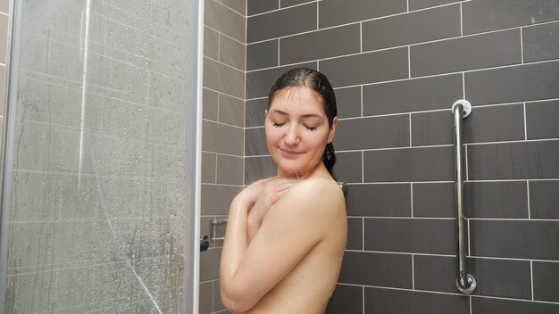 Pretty girl w stands under shower and enjoying in tiled bathroom at home closeup