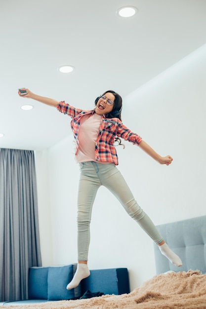 Pretty girl wearing headphones and holding a smartphone while jumping on the bed with her mouth open