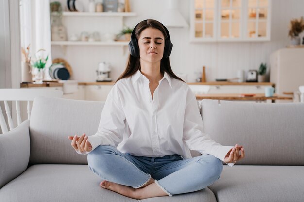 Foto bella ragazza in camicia bianca jeans blu seduto sul divano a casa meditando in cuffia occhi chiusi