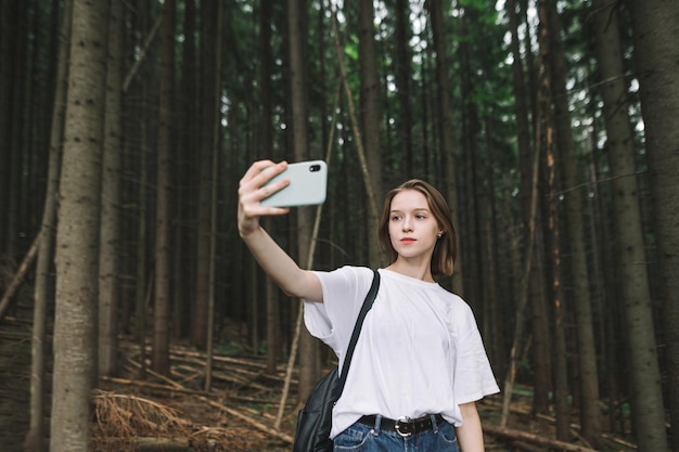 Pretty girl in white tshirt and with backpack makes selfie on smartphone on forest background