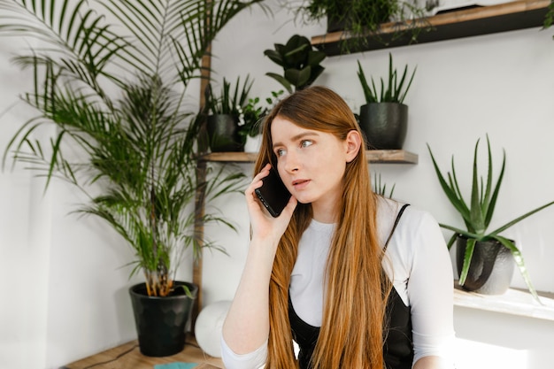 Pretty girl with blond hair talking on the phone indoors with plants and white wall looking away