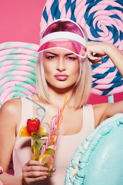 Pretty girl with blonde hair wearing top and pink cap standing with huge sweet lollypops at pink