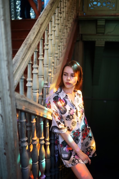 Pretty girl with brown hair in a colorful dress is posing on the stairs