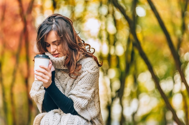 Pretty girl with a cup of hot drink smiles in the forest in the autumn. Woman dressed in a warm sweater