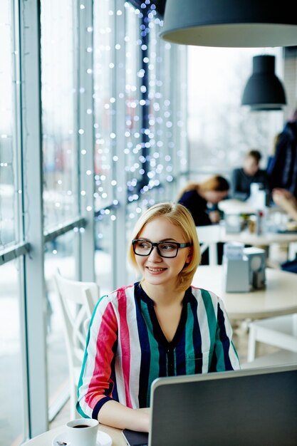 Pretty girl with light hair wearing colorful shirt and glasses sitting in cafe with laptop, freelance concept, online shopping.