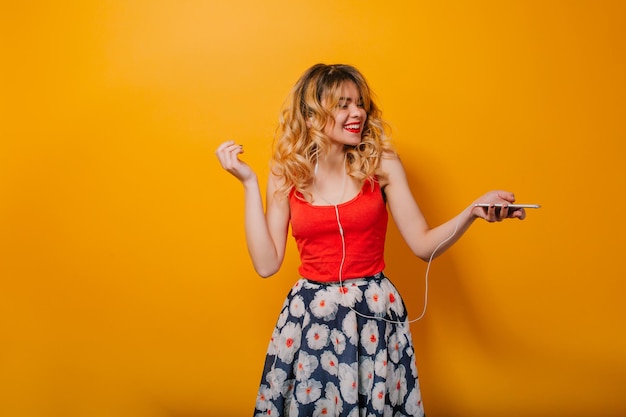 Pretty girl with long curly hair in tail listening to music with headphones on orange background in studio. She wears red T-shirt, skirt flowers, red lips. She is dancing. She looks excited.