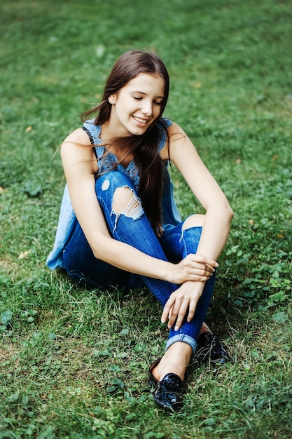 Pretty girl with long hair and torn jeans smiling with closed eyes sitting on grass in park outdoor