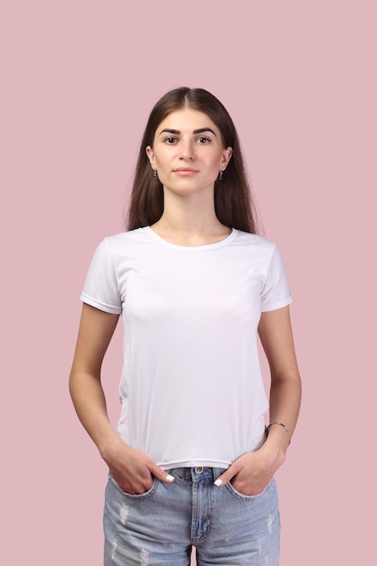 Pretty girl with long hair wearing white T-shirt and posing against light pink background. Adorable brunette looking successful and professional, having positive attitude to life.