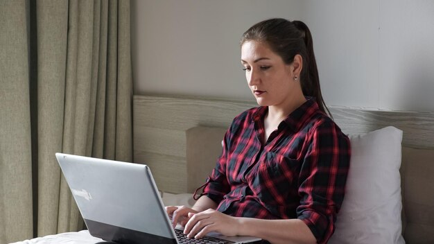 Pretty girl with ponytail in checkered shirt sits on brown bed and types on modern grey laptop