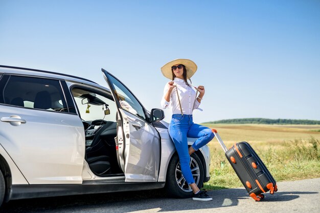 車の近くに立っているスーツケースと彼女の夢の旅のためのwiatを持つかわいい女の子