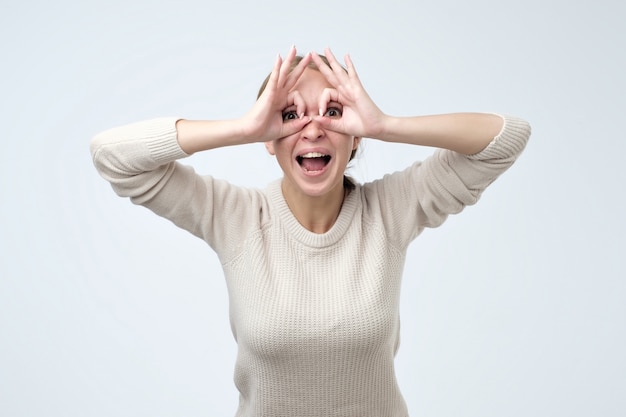 Pretty girl with surprised face holding fingers near eyes like glasses