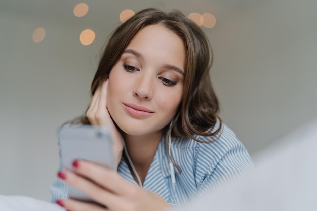 La bella ragazza ama la musica con gli auricolari moderni cerca sui social network sul cellulare ha uno sguardo serio sullo schermo ha una pelle sana trascorre il tempo libero a casa ascolta la trasmissione radiofonica