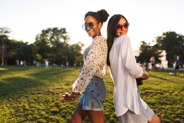 Pretty girls in sunglasses happily looking in camera while playing on little guitar and dancing together in city park