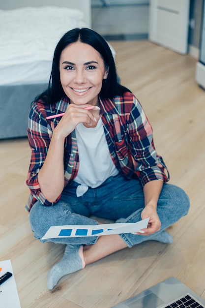 Pretty gladsome young woman in casual clothes working from home\
with a laptop and a graphic chart