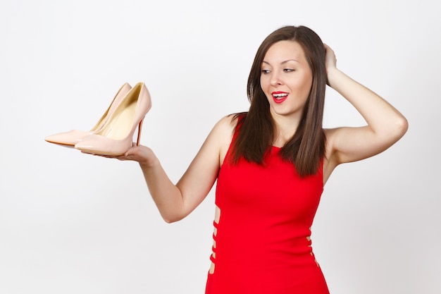 Pretty glamour caucasian fashionable young brown-hair woman in red dress holding beige shoes with red sole of her shopping on palm of hand isolated on white background. Copy space for advertisement.