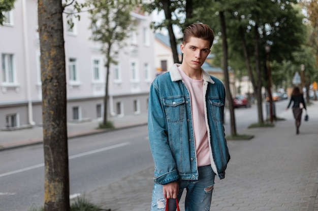 Pretty good-looking young man with stylish hairstyle in fashion pink t-shirt in fashionable blue denim jacket in vintage jeans walks near near road on street. Nice guy in trendy youth clothes enjoy.