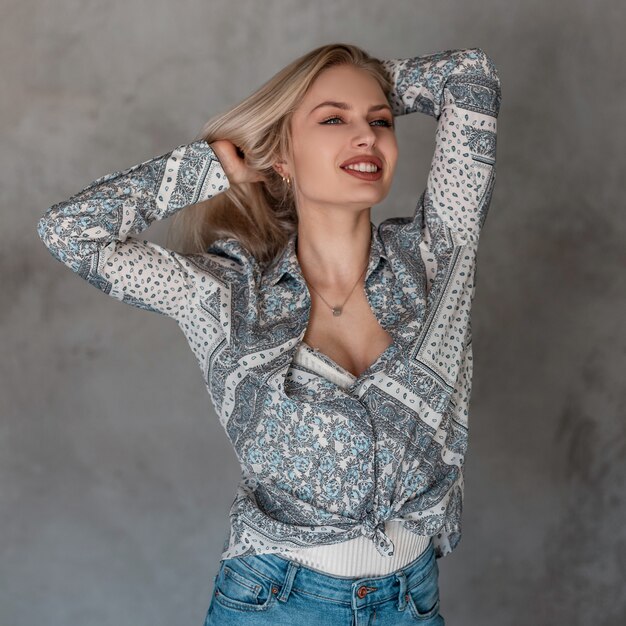 Pretty happy beautiful young blond woman with a cute smile in a vintage gray shirt with a pattern in a white t-shirt in stylish jeans posing in a room near the gray wall