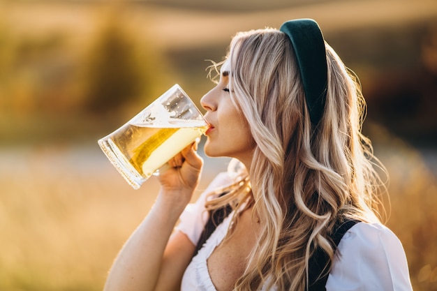 Довольно счастливая блондинка в дирндль, традиционное фестивальное платье, пьет пиво на свежем воздухе в поле