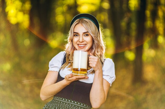 Bionda abbastanza felice in dirndl, abito tradizionale da festa, con boccale di birra all'aperto nella foresta