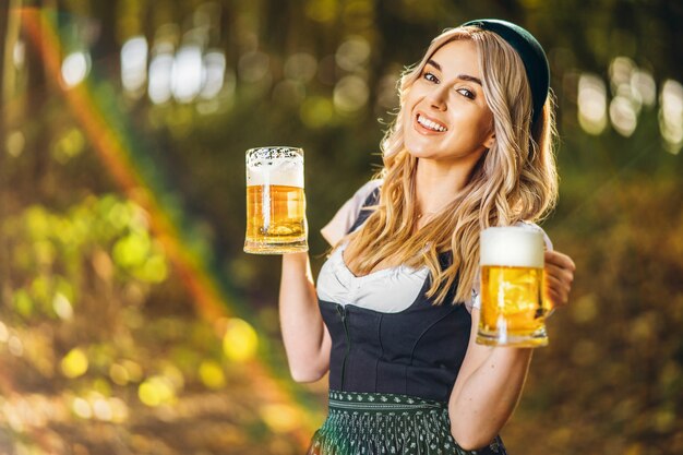 Pretty happy blonde in dirndl, traditional festival dress, holding two mugs of beer outdoors in the forest