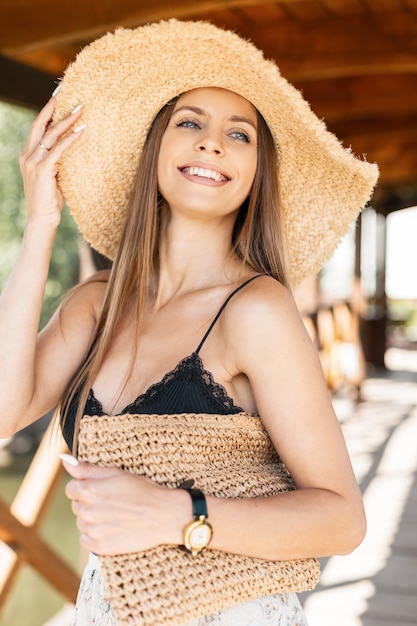 Donna caucasica abbastanza felice con sorriso di bellezza in cappello di paglia di moda con borsa tessuta vintage in top lingerie di pizzo nero di estate che cammina sulla spiaggia durante le vacanze estive