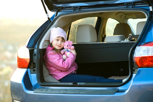 Pretty happy child girl playing with a pink toy teddy bear in a car trunk.