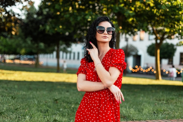 Pretty happy curly woman with retro sunglasses in fashion elegant red dress walks in the city