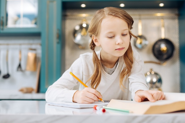 Pretty happy fair-haired girl smiling and writing in her notebook while sitting at the table and reading a book