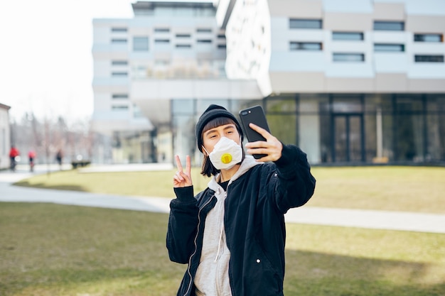 Pretty happy girl in casual wear and with respiratory mask taking selfie on the street while coronavirus pandemic