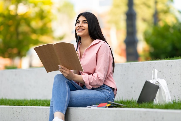 Довольно счастливая латиноамериканка, держащая ручку и пишущая в блокноте, сидящая в парке с мыслями о ноутбуке