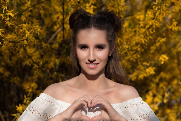 Pretty happy model posing near yellow blooming flowers and showing heart gesture