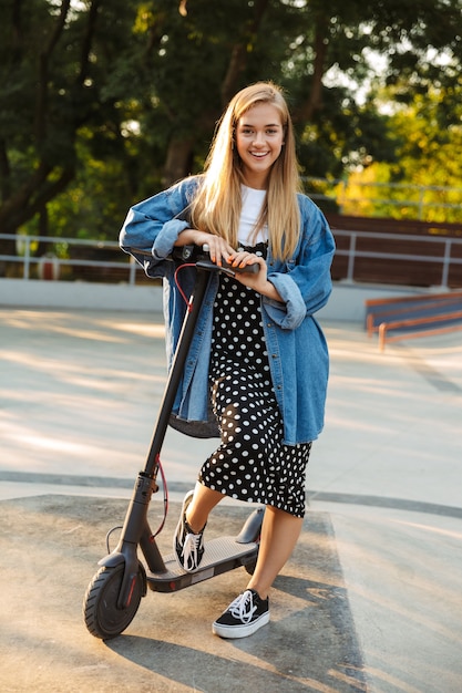 pretty happy optimistic teenage girl in park walking on scooter.